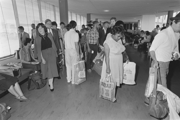 Schwarzes und weißes Bild von Reisenden in einem Flughafen, einige gehen mit Gepäck, andere sitzen auf Bänken oder stehen, mit einem Mann und einer Frau, die Taschen halten, in der Mitte und Fensterjalousien links.