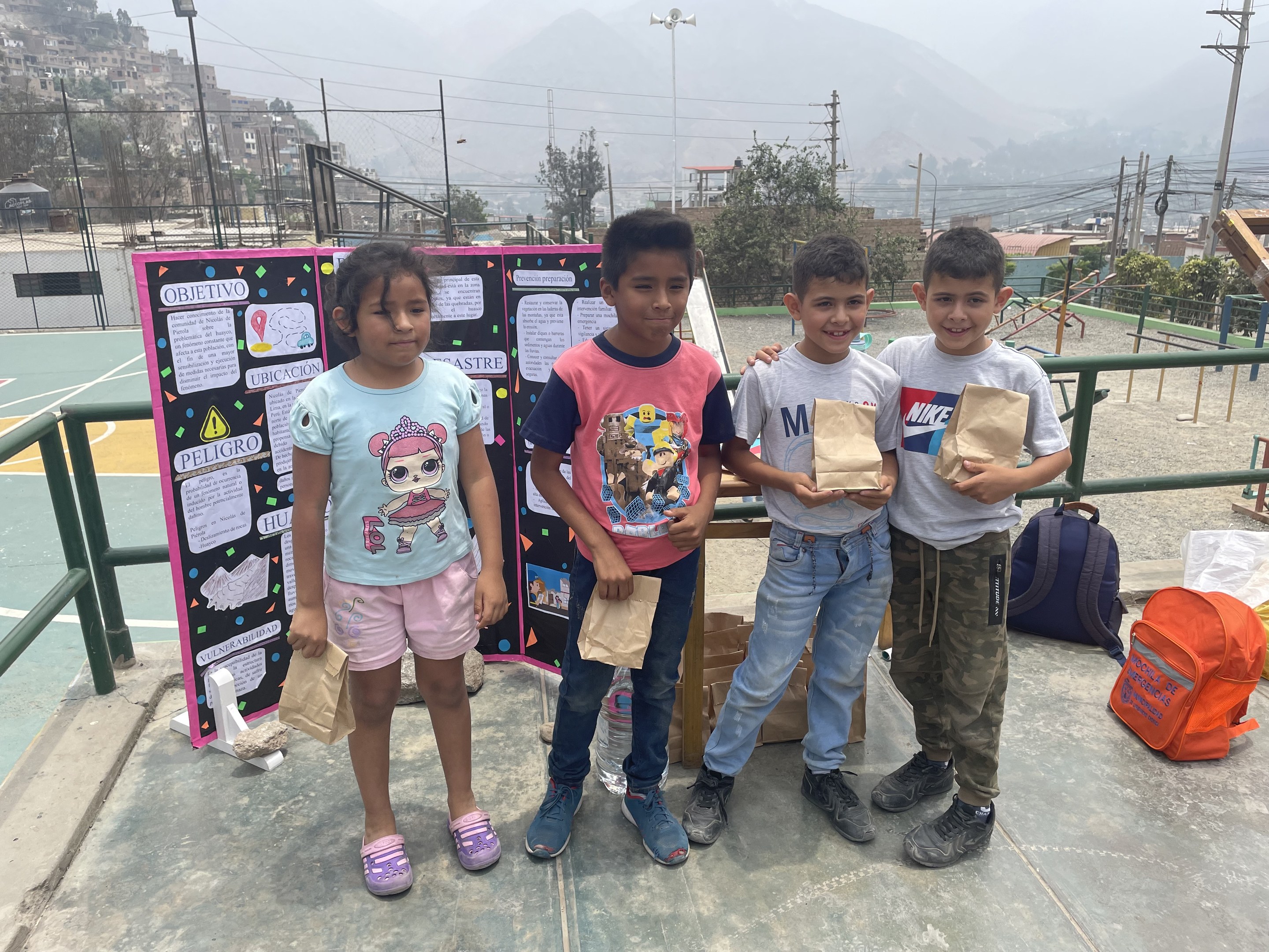 Gruppe von Kindern auf einem Basketballfeld mit Papiertüten in den Händen, mit Gegenständen auf der rechten Seite und einer Tafel mit Text auf der linken Seite, vor einem Hintergrund aus Zäunen, Masten, Straßenlaternen, Bäumen, Gebäuden, Hängen und Himmel.