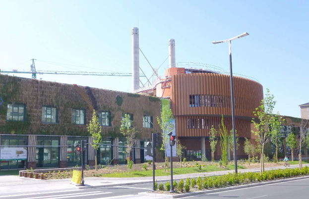 Großes Industriegebäude mit grüner Dachfläche, identifiziert als Battersea Power Station, umgeben von Grünflächen und städtischer Infrastruktur einschließlich Straßenlaternen, Ampeln und Fahrzeugen.