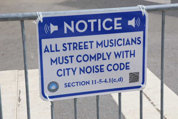 Anschlagtafel an einem Zaun mit einer Tafel, auf der "Anschlag: Alle Straßenmusiker müssen sich an die Stadtlärmvorschriften halten" steht und eine Straße im Hintergrund.