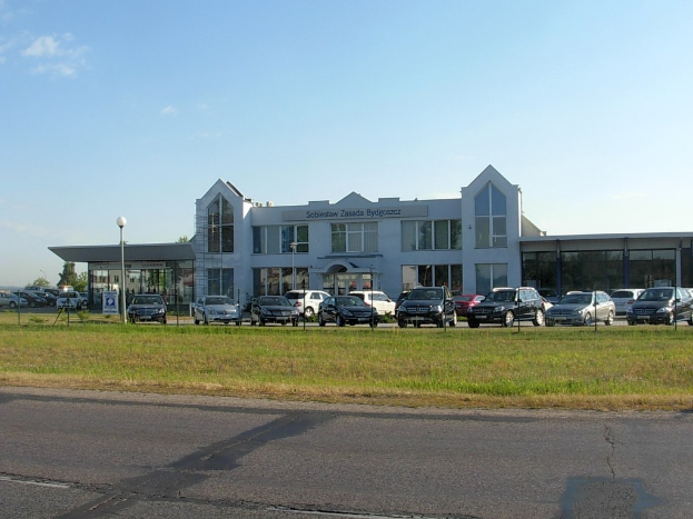 Ein Autohaus mit parkenden Fahrzeugen, umgeben von Gebäuden, Straßeninfrastruktur, fahrenden Autos und Bäumen unter einem bewölkten Himmel.