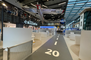 Ein großer Raum mit weißen Wänden, ein vom Himmel hängendes Flugzeug, ein Geländer auf der linken Seite, Gegenstände auf dem Boden auf der rechten Seite, Texttafeln im Hintergrund und an Metallstangen an der Decke befestigte Lampen, wahrscheinlich am Düsseldorfer Flughafen.