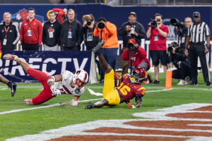 Ein Fußballspieler taucht in die Endzone ein, um einen Touchdown zu erzielen, mit einer Gruppe von Menschen am Boden, einige halten Kameras, und ein Banner mit Text im Vordergrund, während Geländer und Pfosten im Hintergrund sichtbar sind.