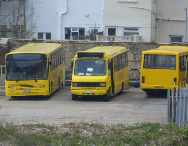 Drei gelbe Busse nebeneinander auf einem eingezäunten Parkplatz mit Grünfläche, einem Gebäude und anderen Strukturen.