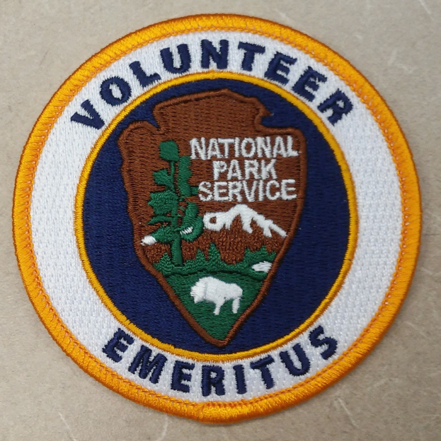 Runder Flicken mit blauem Rand und weißem Hintergrund, der den fetten schwarzen Text "Volunteer National Park Service Emeritus" in der Mitte eines gelben Kreises zeigt.