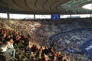 Große Menschenmenge in einem Stadion bei einem Fussballspiel mit einer Bühne, Fahnen, Stangen, einem Bildschirm und der Allianz Arena in München, Deutschland im Hintergrund.