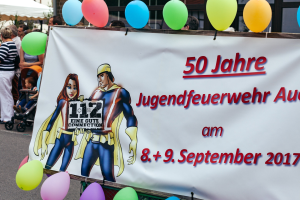 Gruppe von Menschen auf einer Straße mit Kinderwagen, mit einem Banner mit der Aufschrift "50 Jahre Jugendfeuerwehr August 2017", einem Zaun, Luftballons, einem Zelt, einem Gebäude mit Fenstern und Bäumen im Hintergrund.