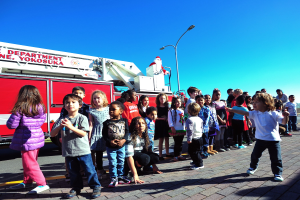 Gruppe von Kindern vor einem Feuerlöschfahrzeug, das als Teil des Weihnachtsparades der Yokosuka-Feuerwehr auf einer Straße mit Laternenpfählen, Bäumen und einem klaren blauen Himmel steht.