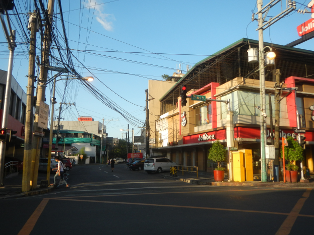 Eine Stadtstraße mit Autos, Fußgängern, Strommasten mit Drähten, Laternen, Ampeln, Gebäuden mit Schildern, Topfpflanzen und einer bewölkten Himmel.