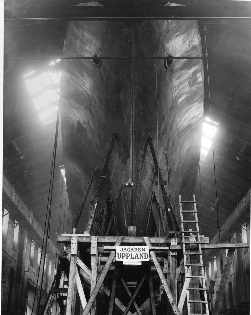 Großes schwarzes und weißes Schiff in Bau in einer Werft mit Stützkonstruktionen, Leiter, Schild, Deckenleuchten und Fenstern.