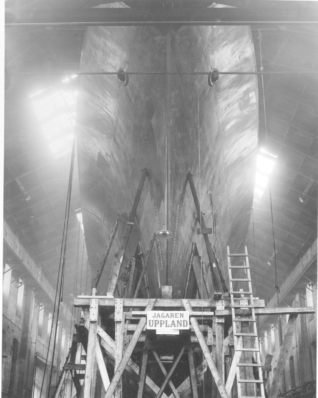 Großes schwarzes und weißes Schiff in Bau in einer Werft mit Stützkonstruktionen, Leiter, Schild, Deckenleuchten und Fenstern.