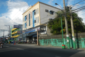 Stadtstraße mit Gebäuden, Strommasten, Kabeln, Straßenlaternen, Fahrzeugen, Schildern, Bäumen und einem bewölkten Himmel, mit einem Gebäude im Vordergrund, das dicht mit Drähten bedeckt ist.