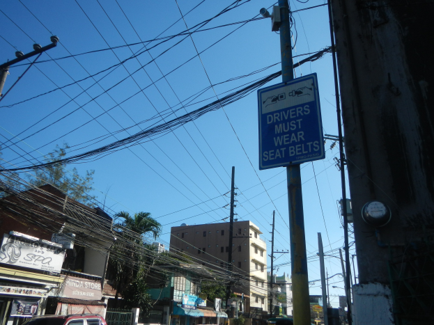 Stadtstraße mit Fahrzeugen, Strommasten, Gebäuden, Bäumen und einem "Fahrer müssen den Sicherheitsgurt anlegen"-Schild an einem Pfahl, unter einem sichtbaren Himmel.