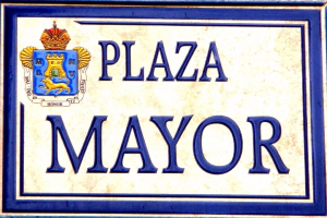 Ein rechteckiges weißes Schild mit schwarzer Schrift, die "Plaza Mayor" liest und einen blauen Rahmen hat, mit einem gekrönten Wappen-Logo darauf, das an einem Gebäude angebracht ist.