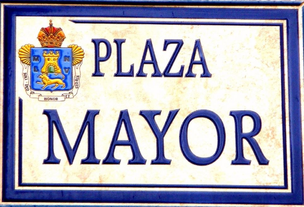Ein rechteckiges weißes Schild mit schwarzer Schrift, die "Plaza Mayor" liest und einen blauen Rahmen hat, mit einem gekrönten Wappen-Logo darauf, das an einem Gebäude angebracht ist.
