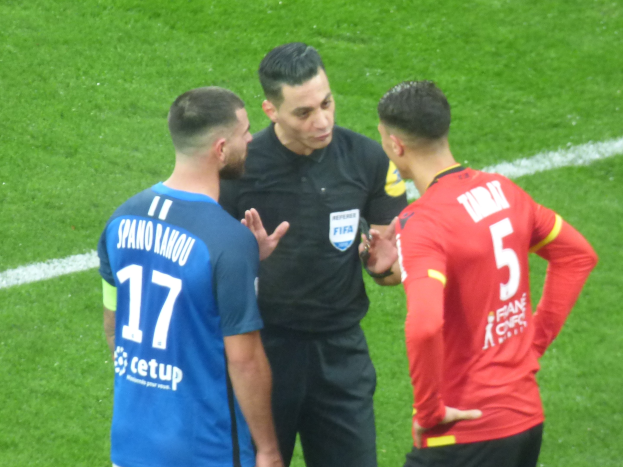 Schiedsrichter in schwarzer Kleidung spricht mit zwei Männern auf einem Fußballfeld, einer in blau-roten T-Shirts mit Text und Brille, der andere ebenfalls mit Brille.