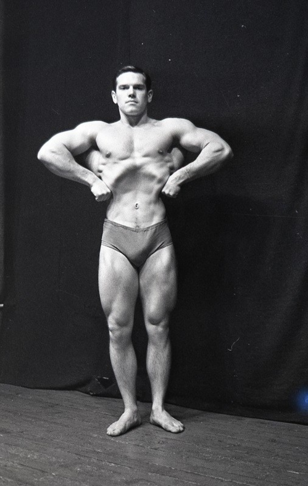 Bodybuilder auf einer Bühne mit händen an den hüften, in schwarz-weißer ausrüstung, mit einem Vorhang im Hintergrund.