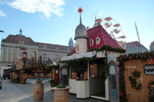 Ein belebter Weihnachtsmarkt in einer Stadtmitte mit geschmückten Bäumen, Gebäuden, Fahrzeugen, Passanten und Wolkenhimmel.