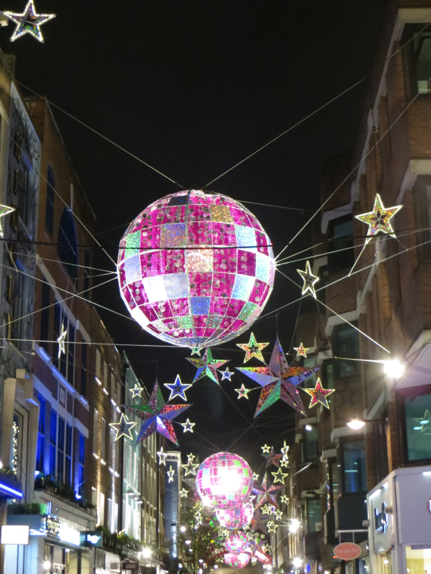 Eine Stadtstraße mit Lichtern und einer großen Diskokugel, die von einem Gebäude hängt, mit zusätzlichen Lichtern an Strukturen, unter einem dunklen Himmel.