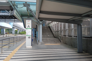 Ein Bahnsteig mit Treppen, eine Straße mit Geländern auf der linken Seite und ein Hintergrund mit Bäumen, Pfählen, Gebäuden und einer Brücke; oben auf dem Bahnsteig gibt es Rohre und eine Tafel mit Text.
