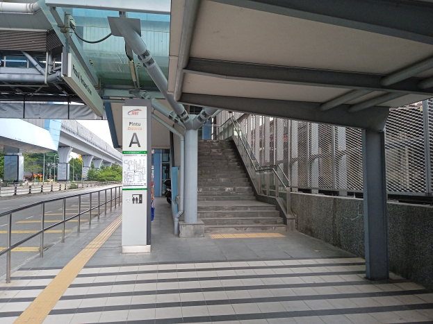 Ein Bahnsteig mit Treppen, eine Straße mit Geländern auf der linken Seite und ein Hintergrund mit Bäumen, Pfählen, Gebäuden und einer Brücke; oben auf dem Bahnsteig gibt es Rohre und eine Tafel mit Text.