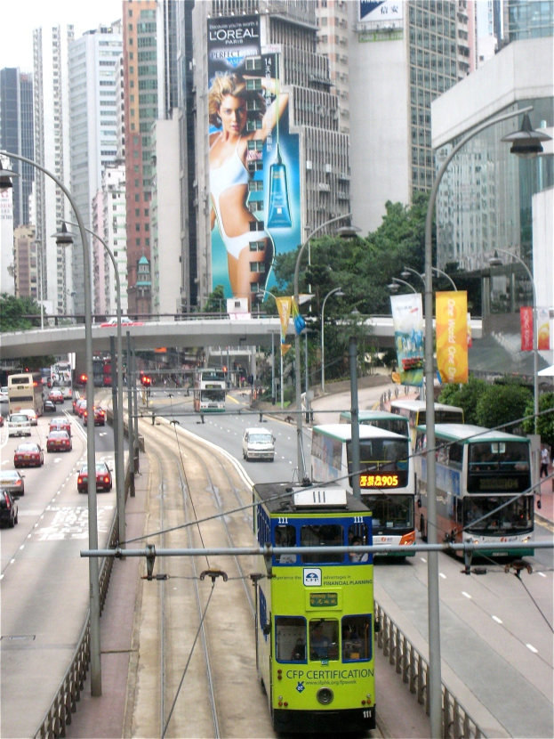 Eine belebte Stadtstraße mit Doppeldeckerbussen, Autos und anderen Fahrzeugen, umgeben von hohen Gebäuden, Bäumen und einer Brücke unter einem klaren blauen Himmel, mit Straßenschildern und Bannern.