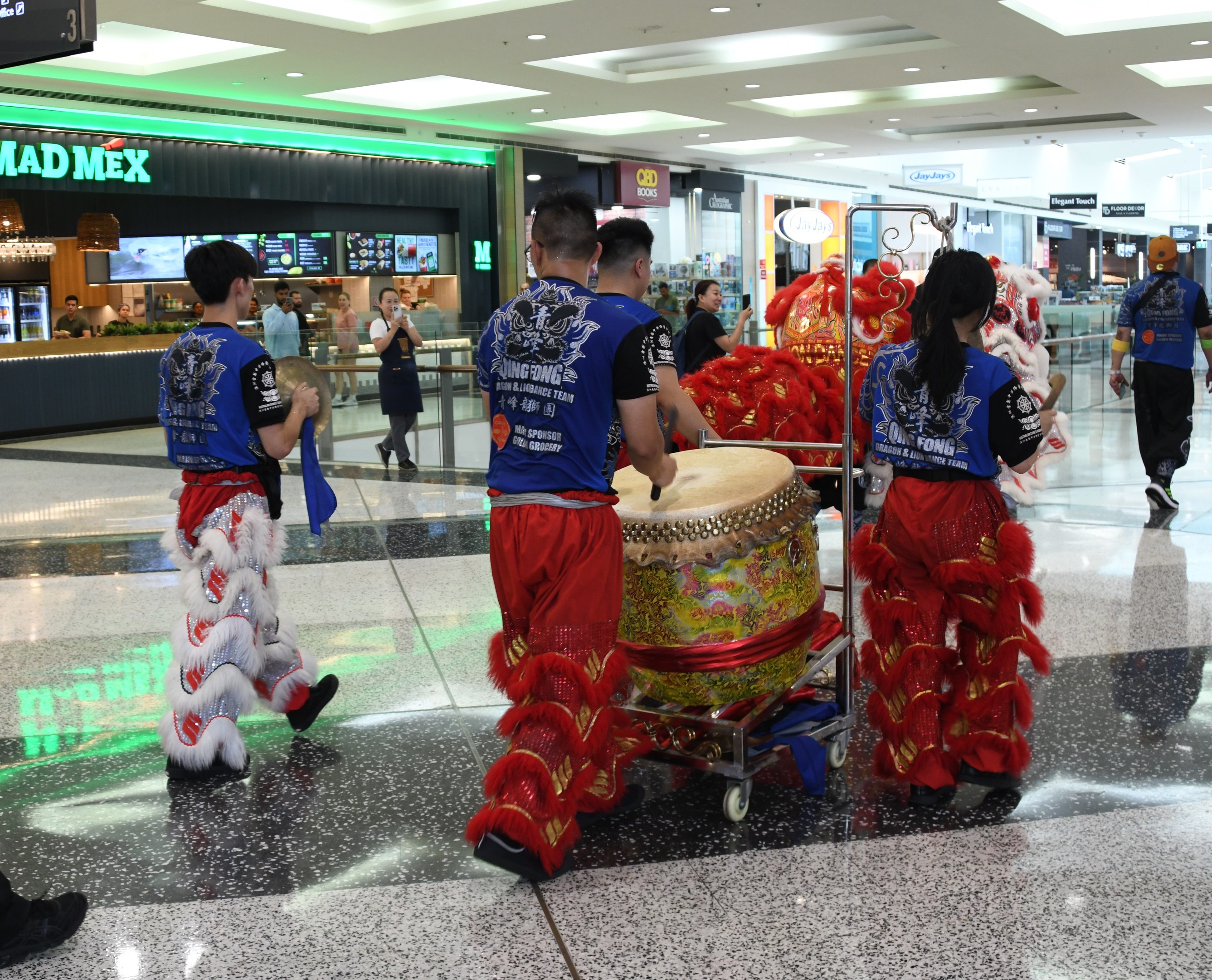 Eine Gruppe von Menschen in roter und blauer Kleidung mit Musikinstrumenten und Wagen durchquert einen hell erleuchteten Flughafen w├Ąhrend einer Feierlichkeit, mit Gesch├Ąften und Schildern im Hintergrund.