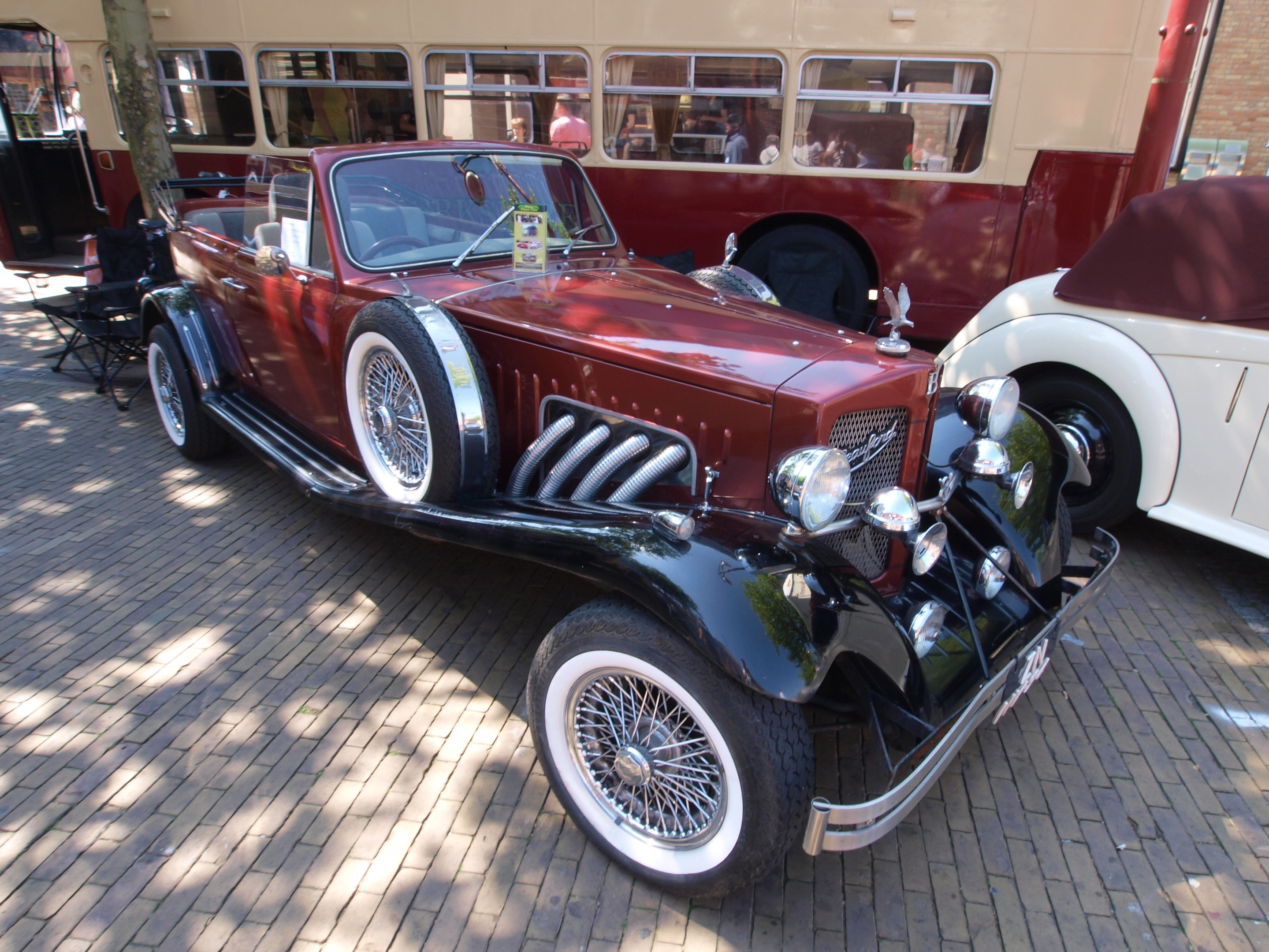 Ein Vintage-Auto neben einem Doppeldeckerbus auf einer Kopfsteinpflasterstraße geparkt, mit Stühlen und einem Baum im Vordergrund, einem Gebäude im Hintergrund und Menschen im Bus sichtbar.