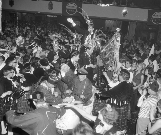 Ein Schwarz-Weiß-Bild einer Menge von Menschen in Schottenröcken, die auf einer Party tanzen, wobei einige Musikinstrumente und Texttafeln im Hintergrund halten.