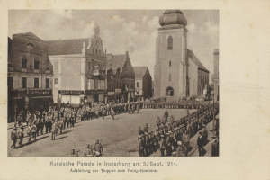 Ein Schwarz-Weiß-Foto eines Umzugs in Insterburg am 5. September 1914 mit vielen Menschen, Gebäuden im Hintergrund und Text am unteren Bildrand.