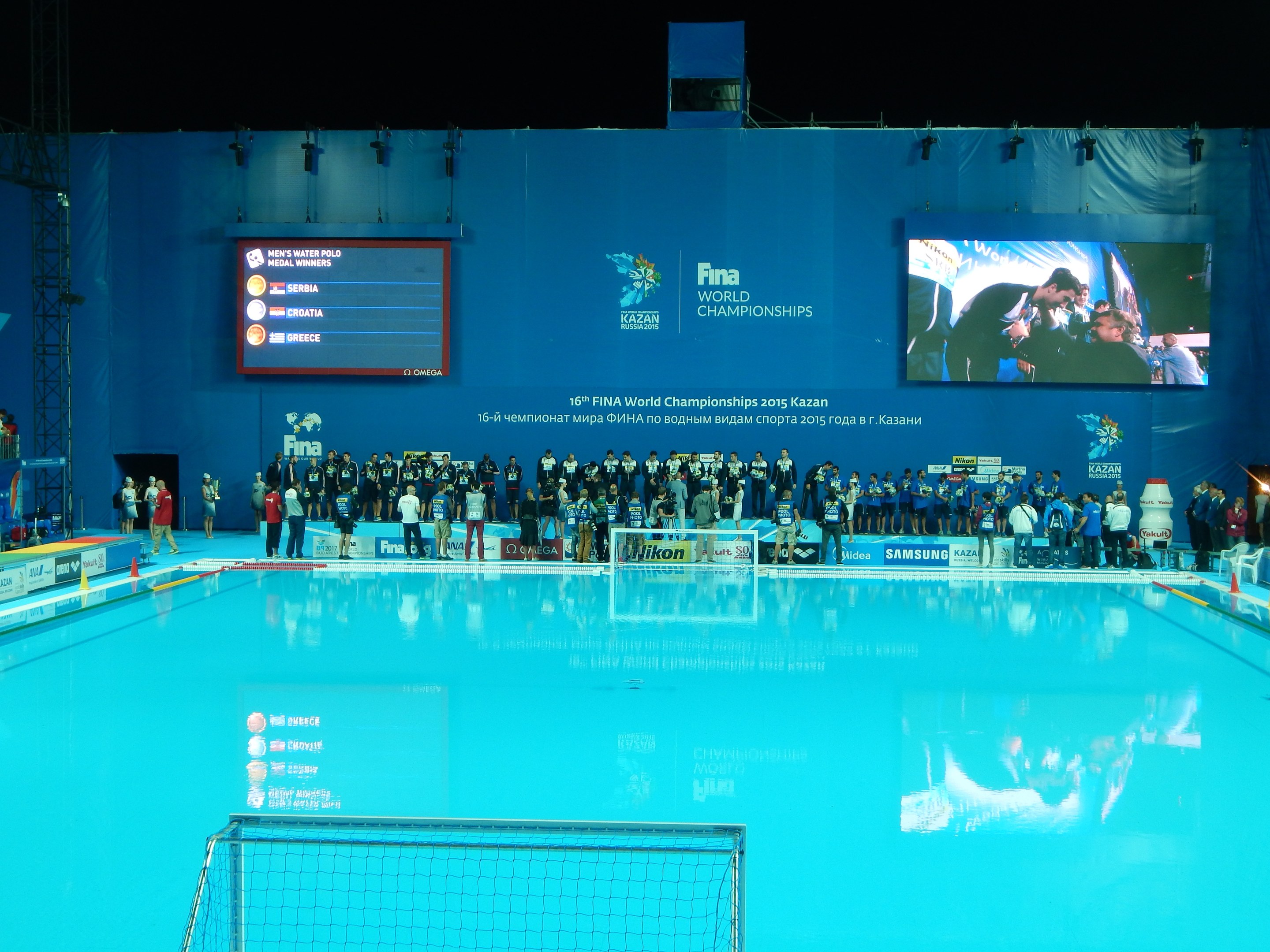 Gruppe von Menschen um ein Hallenbad mit Netz am Boden herumstehend, mit einer Wand im Hintergrund, die "FINA World Championships 2015 Kazan" auf einem Bildschirm anzeigt, zusammen mit Trägern und Lampen.