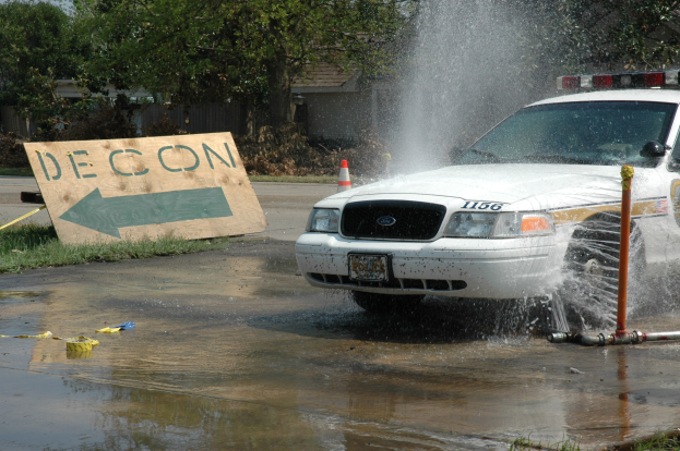 Polizeifahrzeug fährt durch eine Pfütze mit verstreuten Gegenständen, darunter eine Tafel mit Text und ein Verkehrskegel, Bäume und Gebäude im Hintergrund unter einem klaren blauen Himmel.