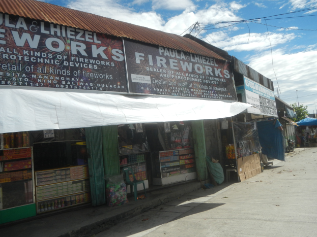 Ein Geschäft mit einer 'Feuerwerk' -Schilder, umgeben von verschiedenen Gegenständen und anderen Geschäften, mit Bäumen, Pfählen, Drähten und einem bewölkten Himmel im Hintergrund.