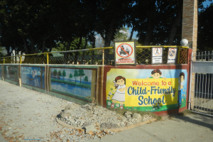 Ein Wandschild mit der Aufschrift "Willkommen in einer kinderfreundlichen Schule" vor einem Metallzaun, einer Säule, Bäumen, Steinen und einem Weg.