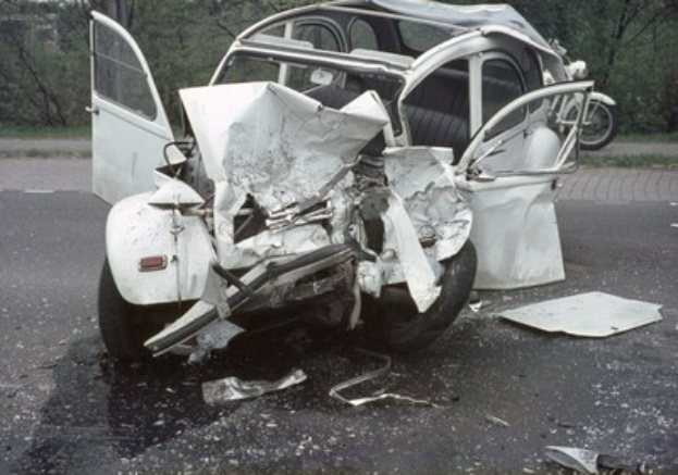 Schwer beschädigte Limousine am Straßenrand mit Frontalcrash, umgeben von Bäumen und einem entfernten Gebäude.