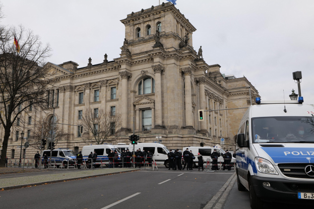 Eine Gruppe von Polizisten steht vor dem Reichstaggebäude in Berlin, Deutschland, mit Fahrzeugen, einem Zaun, Verkehrsampeln, Laternenpfählen, Bäumen und Flaggen im Hintergrund, unter einem klaren Himmel.