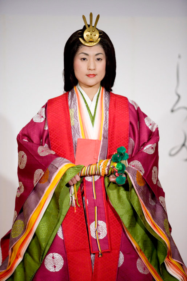 Eine Frau in einem traditionellen japanischen Kimono, das ein leuchtendes rotes und grünes Kleid mit aufwändigen Mustern zeigt, steht selbstbewusst auf einem Laufsteg, ihr Haar zu einem ordentlichen Knoten gebunden.