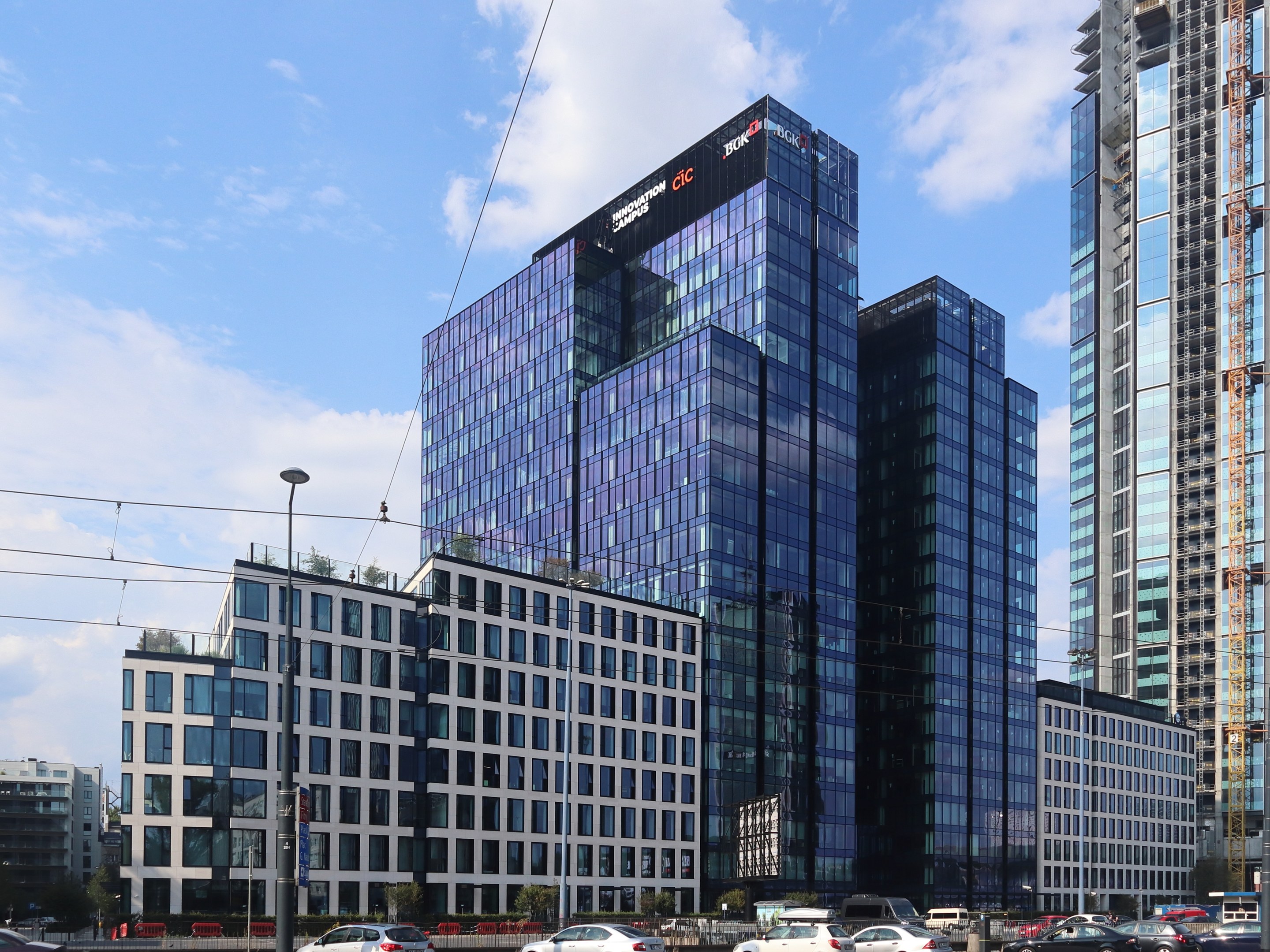 Zentrale der Deutschen Bank in Frankfurt, Deutschland, ein großes mehrstöckiges Gebäude mit zahlreichen Fenstern, umgeben von städtischer Infrastruktur einschließlich Straßenlaternen, Fahrzeugen, Grünflächen und einem bewölkten Himmel.