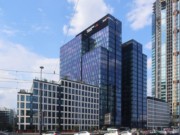 Zentrale der Deutschen Bank in Frankfurt, Deutschland, ein großes mehrstöckiges Gebäude mit zahlreichen Fenstern, umgeben von städtischer Infrastruktur einschließlich Straßenlaternen, Fahrzeugen, Grünflächen und einem bewölkten Himmel.