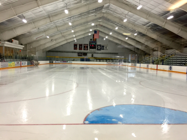 Großes Halleneisstadion mit Deckenbeleuchtung, Werbetafeln, einer Rückwand, Treppen mit Geländern und einigen Menschen, was auf eine Sportanlage hinweist, die likely für Hockey genutzt wird.