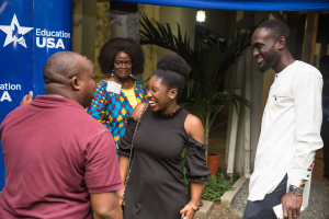 Gruppe von Menschen, die vor einem blauen Zelt mit einem Banner, Topfpflanzen, einer Säule, Wand und einer Decke mit Leuchten sprechen.