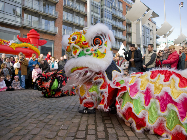Ein lebendiges chinesisches Neujahrsfest in Amsterdam mit einer Löwen-Tanzvorstellung vor einer Zuschauermenge, darunter einige, die das Ereignis fotografieren, vor einer Kulisse aus Gebäuden, Laternenmasten und einem klaren blauen Himmel.