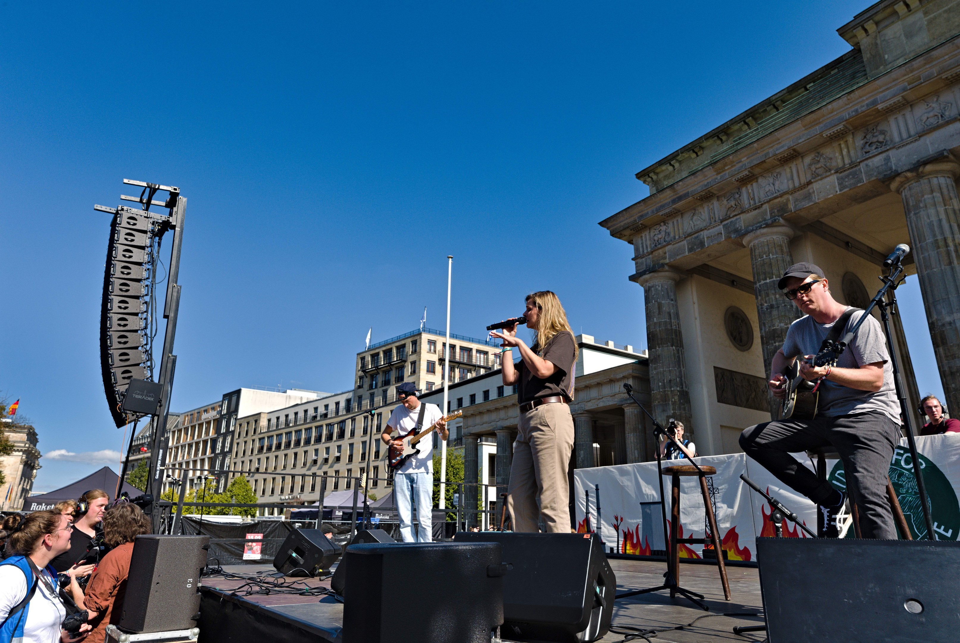 Eine Gruppe von Menschen, die auf einer Bühne Musik spielen, mit dem Brandenburger Tor im Hintergrund, begleitet von Lautsprechern und Equipment unter einem klaren blauen Himmel.