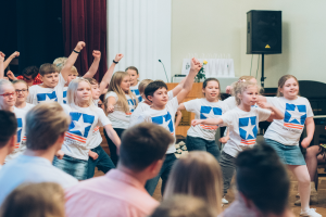Eine Gruppe von Kindern in weißen Hemden und blauen Shorts tanzt auf einer Bühne vor einem sitzenden Publikum, mit einem Vorhang, Lautsprecher und einem Tisch mit Mikrofon im Hintergrund.