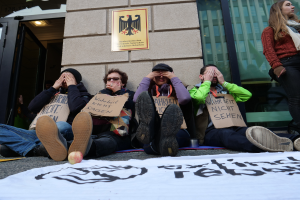 Eine Gruppe von Menschen sitzt vor einem Gebäude auf dem Boden und hält Schilder, mit einer Fahne und einem Apfel auf der Straße, und eine Frau steht rechts daneben und sieht besorgt aus aufgrund einer Regierungsentscheidung.