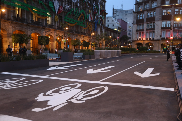 Stadtstraße mit einem ausgewiesenen Fahrradweg, umgeben von Gebäuden und Straßenmöblierung, Menschen auf dem Gehweg und einem klaren blauen Himmel.