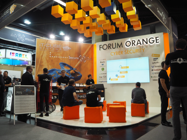 Eine Gruppe von Menschen an einem Messestand für IT-Cube Systems Forum Orange Cyber Security, einige sitzen und andere stehen, in der Nähe einer Tafel, eines Bildschirms, Regalen und Schildern.