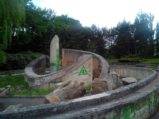 Abandonierter Skatepark mit graffiti-bedeckten Wänden, umgeben von Felsen, Gras, Pflanzen und Bäumen unter einem klaren blauen Himmel.