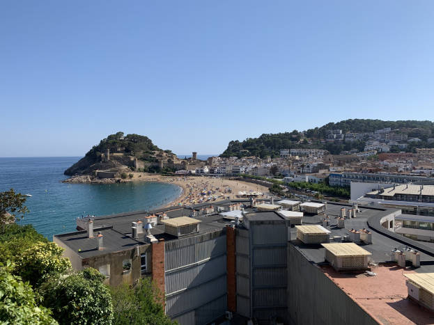 Luftaufnahme eines Strandes in Costa Brava, Spanien, der den Ozean auf der linken Seite, Bäume und Gebäude im Vordergrund und einen hügeligen Wald im Hintergrund zeigt.
