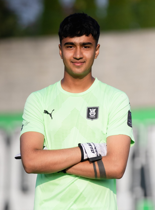 Ein junger Mann in einem grünen T-Shirt steht mit verschränkten Armen und einem Lächeln auf einem Fußballfeld, mit einem unscharfen Hintergrund.